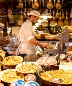 Shopkeeper at a traditional Bologna market stall surrounded by local delicacies like fresh pasta, cheeses, and prosciutti