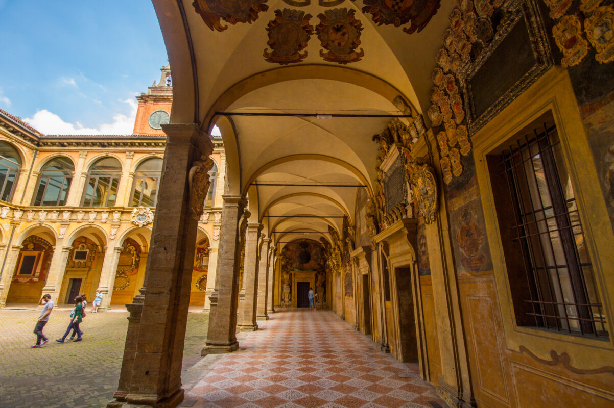 Exterior of the Archiginnasio Palace in Bologna, seen from the street with people under the arcades.